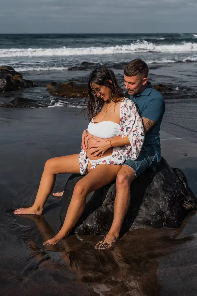 Werdendes Paar sitzt auf einem Felsen – Hände auf dem Babybauch am Playa del Inglés