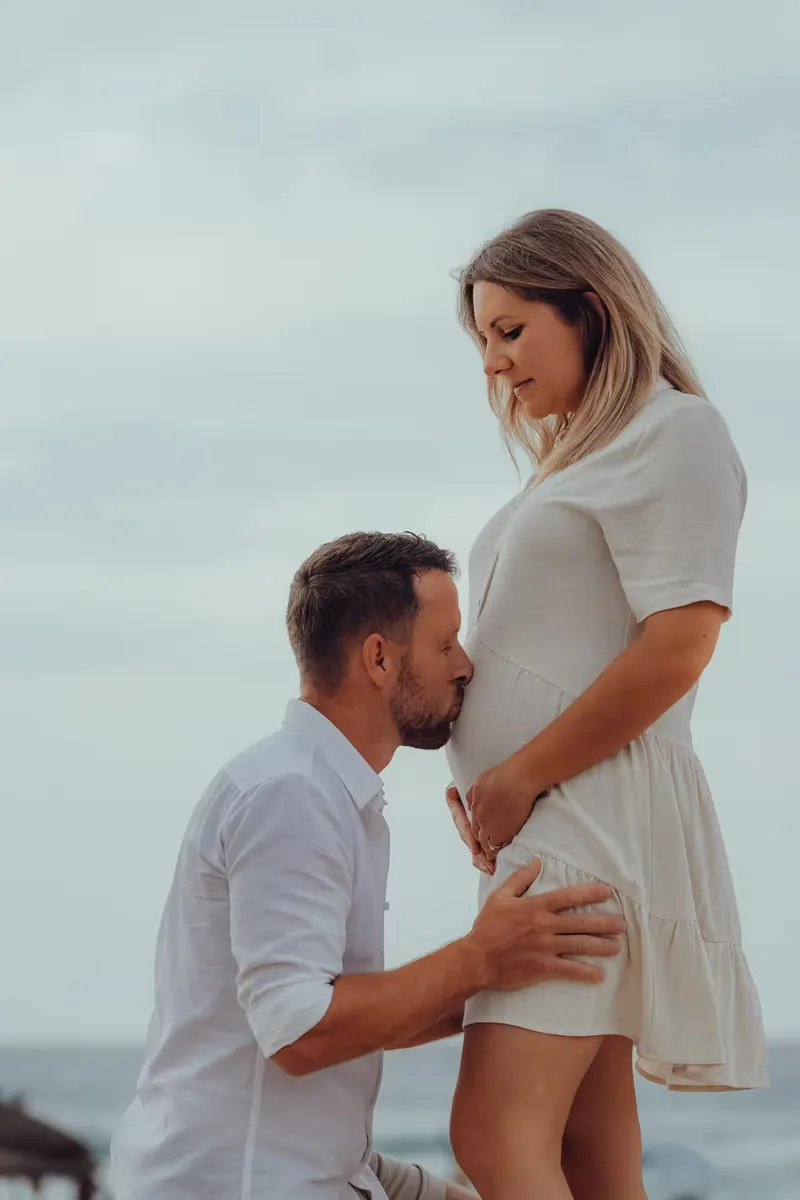 Expectant mother in flowing white dress walking along the shoreline in Mallorca
