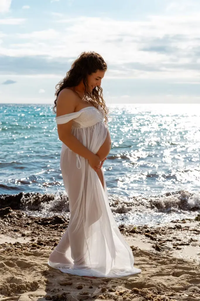 Frau mit Babybauch im weißen durchsichtigen Kleid steht am Meer von Cala Millor