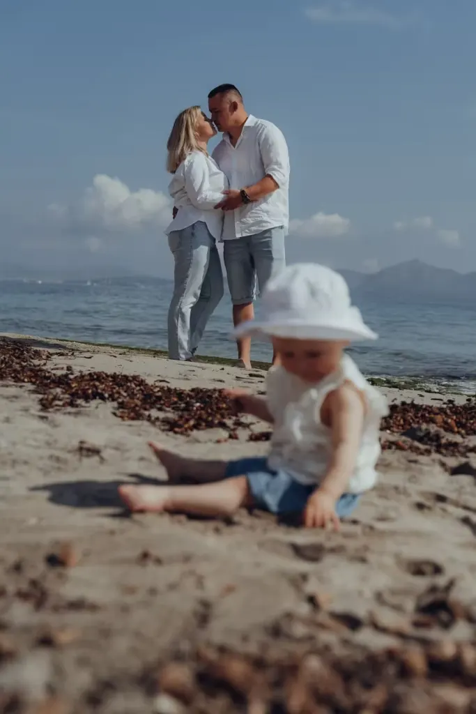 Baby sitzt im Sand während die Eltern sich im Hintergrund küssen am Meer