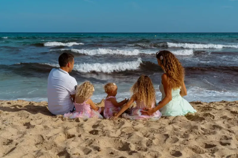 Fünfköpfige Familie mit drei Töchtern sitzt von hinten im Sand und blickt aufs Meer