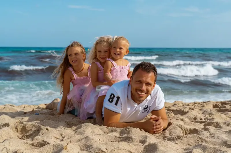 Glücklicher Vater liegt im Sand während seine drei Töchter lachend auf seinem Rücken sitzen