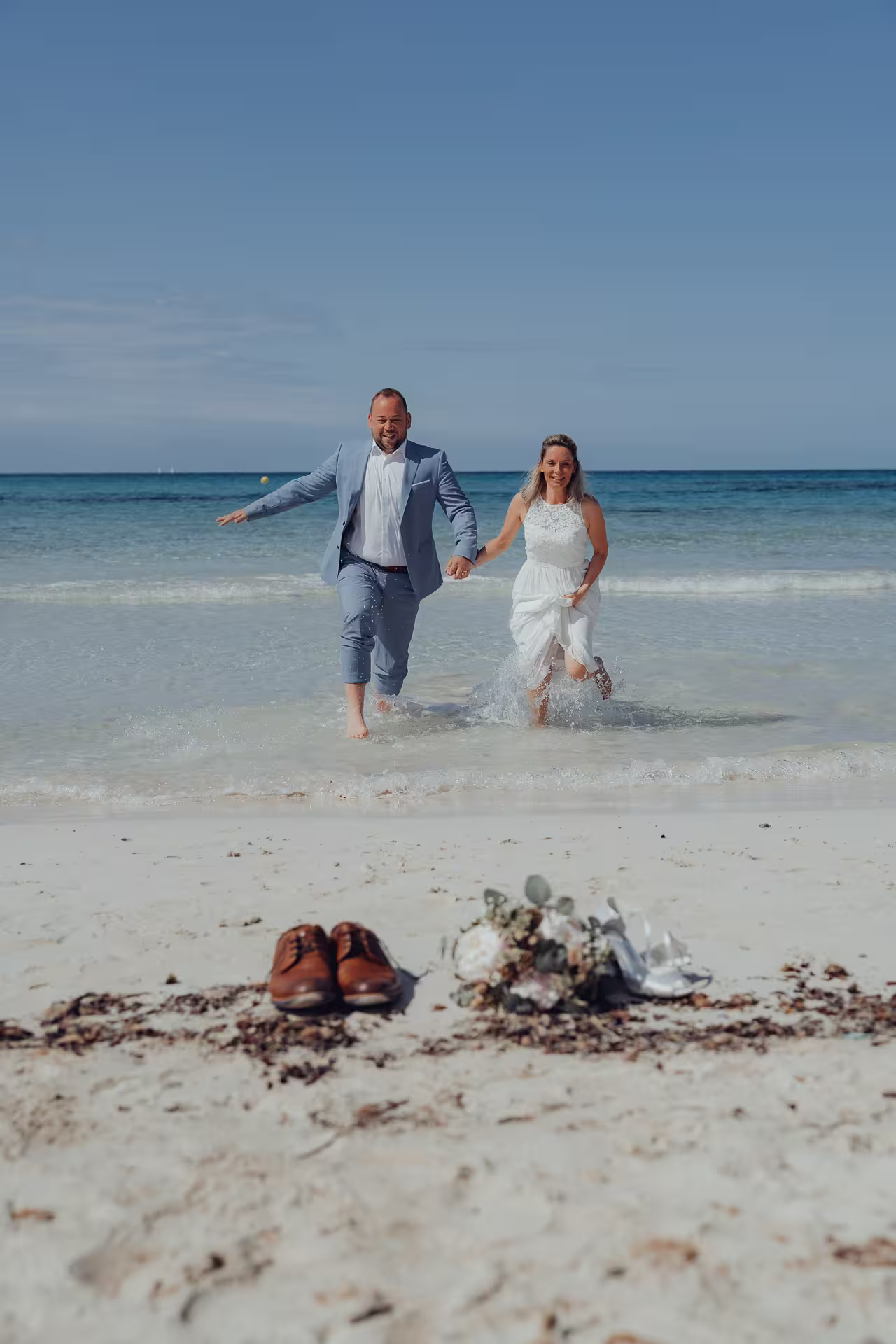 Fotoshooting After Wedding Mallorca: Ein glückliches Brautpaar rennt voller Freude und Händchen haltend durch das flache Wasser am Strand.