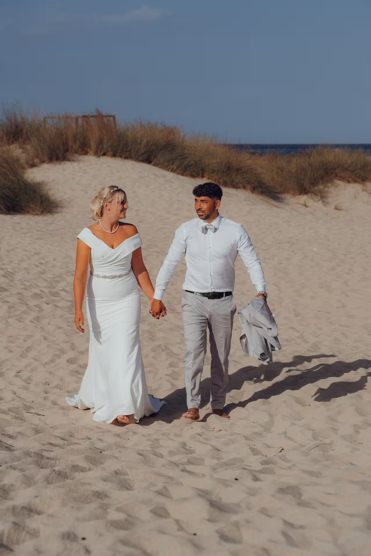 Fotoshooting After Wedding Mallorca: Ein Brautpaar spaziert bei Sonnenuntergang Hand in Hand durch die Sanddünen am Meer.