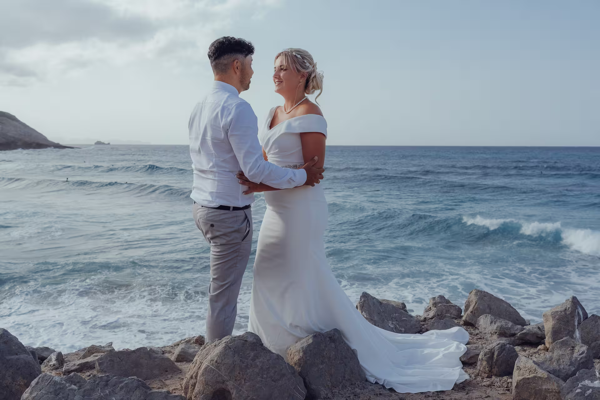 Fotoshooting After Wedding Mallorca: Ein Brautpaar umarmt sich innig an einer Felsküste und blickt dabei auf das aufgewühlte Meer.