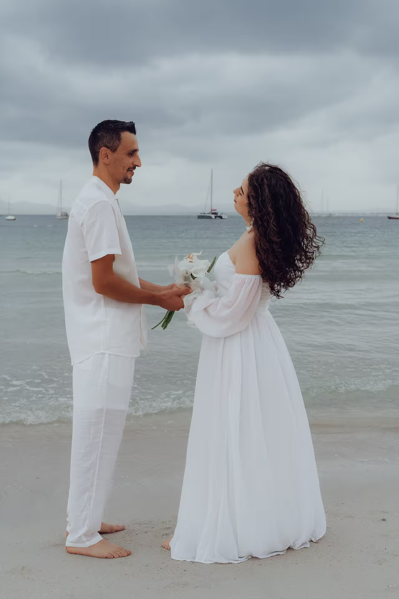 Fotoshooting After Wedding Mallorca: Ein Brautpaar ganz in Weiß steht sich an einem Strand gegenüber, im Hintergrund liegen Segelboote vor Anker.