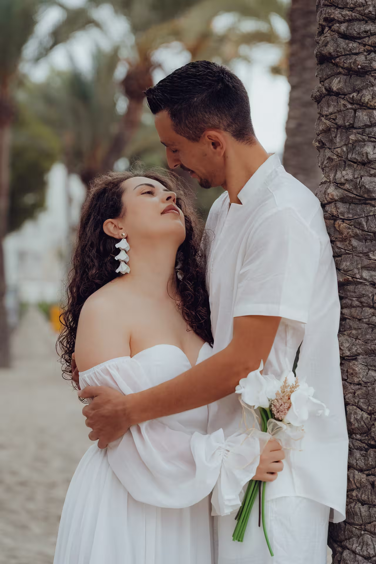 Fotoshooting After Wedding Mallorca: Intimes Porträt eines Brautpaares, das sich an den Stamm einer Palme lehnt und sich liebevoll ansieht.