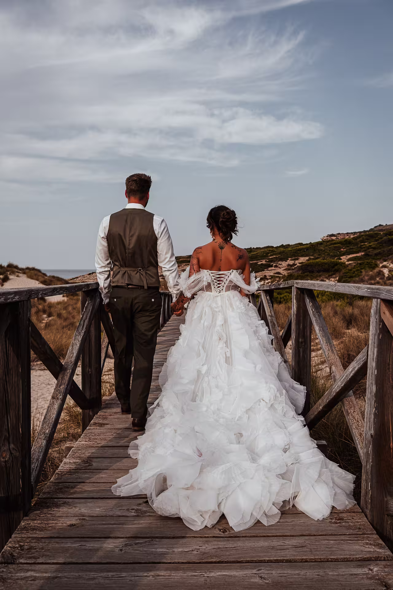 Fotoshooting After Wedding Mallorca: Das Brautpaar spaziert von hinten gesehen Hand in Hand über einen langen Holzsteg durch die Dünen.