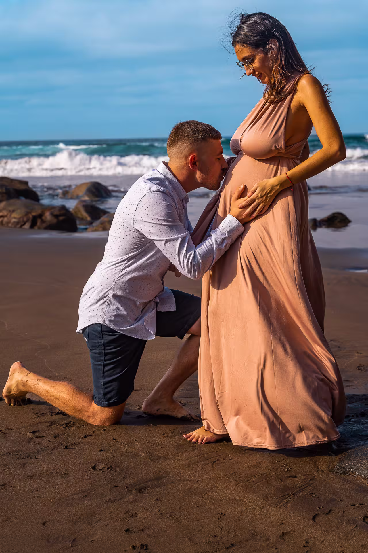 Der werdende Vater küsst zärtlich den Babybauch seiner Partnerin am dunklen Sandstrand von Gran Canaria. Romantisches Babybauch Fotoshooting Gran Canaria mit Meeresrauschen