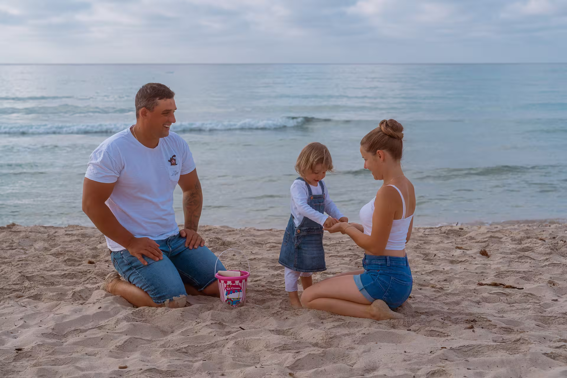 Mallorca Urlaubsfotoshooting Familie: Eltern knien mit ihrer kleinen Tochter im Sand am Strand und spielen gemeinsam am Meer.