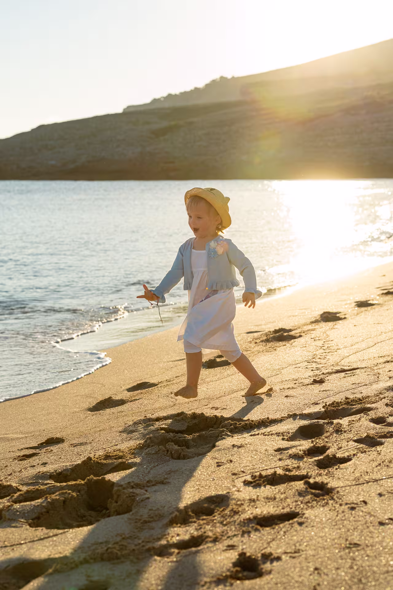 Mallorca Urlaubsfotoshooting Familie: Ein kleines Mädchen mit Strohhut rennt fröhlich bei Sonnenuntergang am Strand entlang des Wassers.