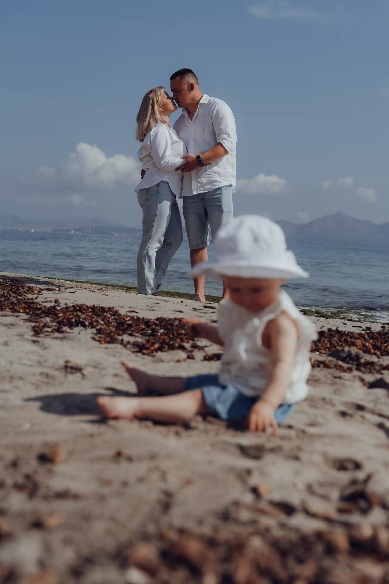 Mallorca Urlaubsfotoshooting Familie: Ein Baby sitzt im Sand am Strand, während die Eltern im Hintergrund unscharf am Meer stehen und sich küssen.