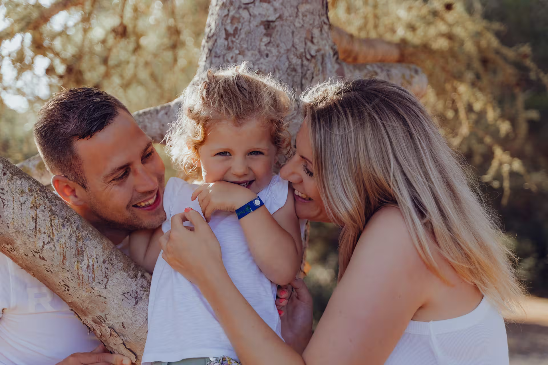 Mallorca Urlaubsfotoshooting Familie: Eltern und ihre kleine Tochter lachen herzlich zusammen und lehnen in warmem Licht an einem Pinienbaum.