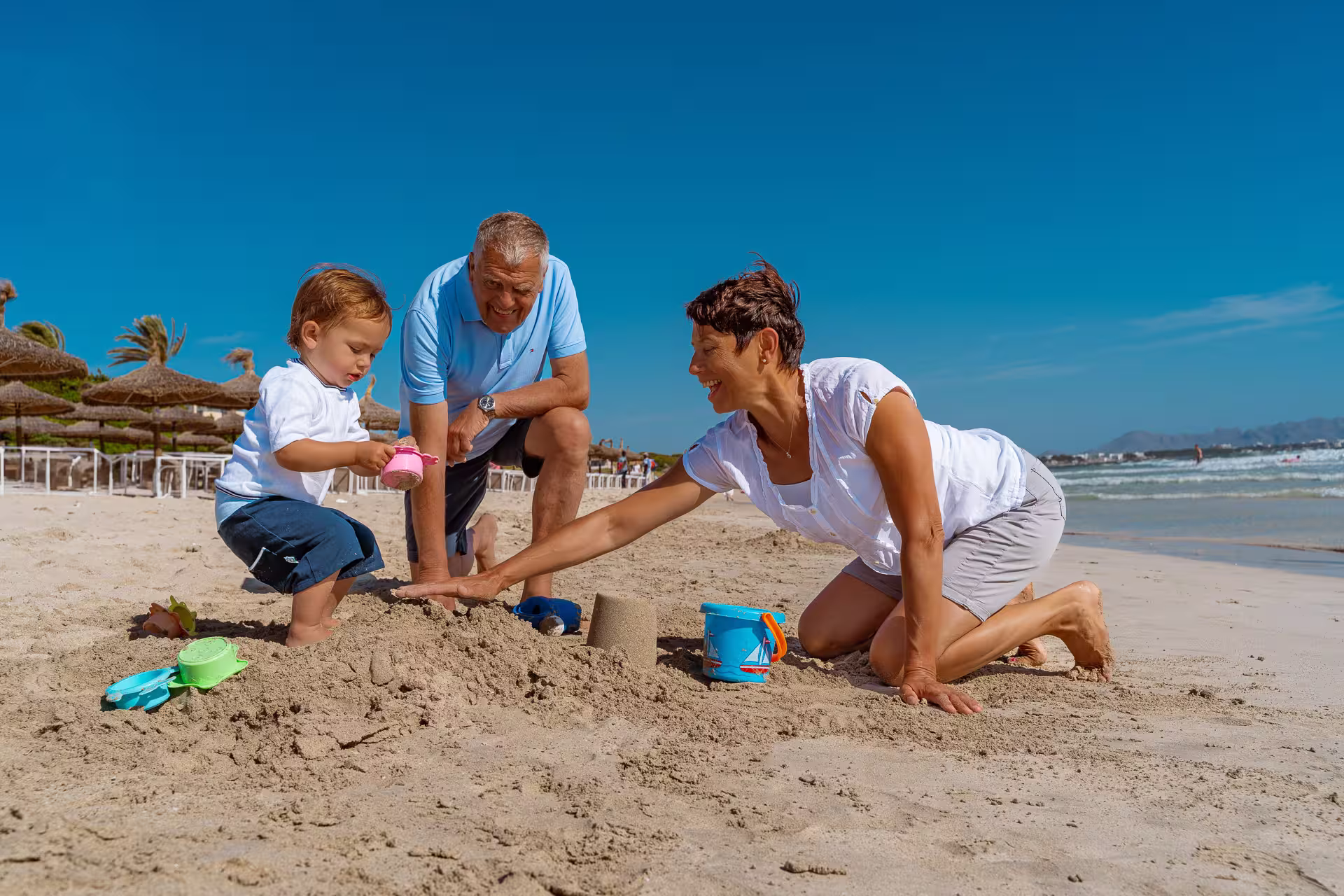 Mallorca Urlaubsfotoshooting Familie: Großeltern bauen mit ihrem kleinen Enkelsohn eine Sandburg am sonnigen Strand von Mallorca.