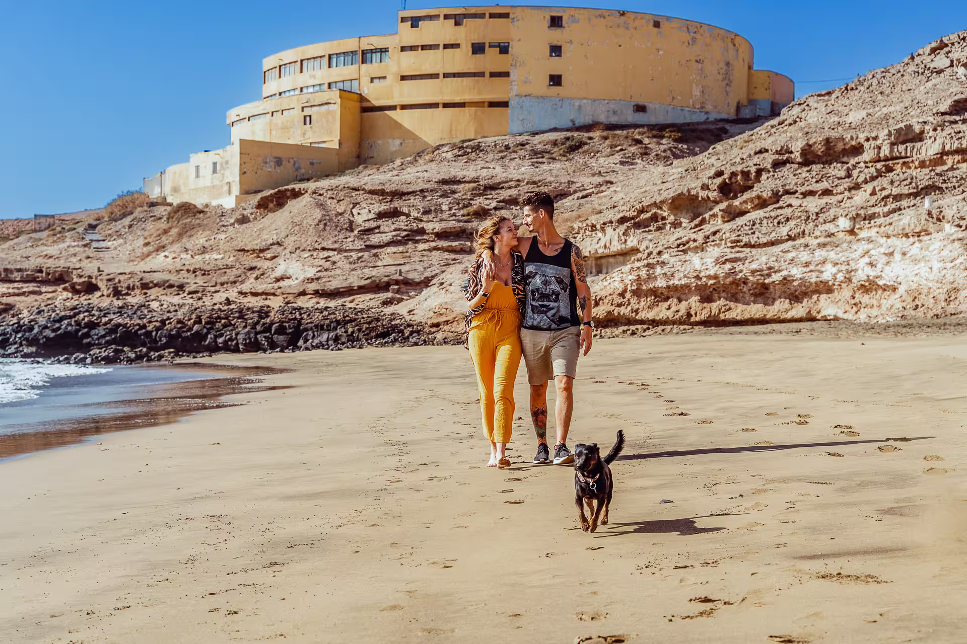 unges Paar mit Hund spaziert entspannt am Sandstrand entlang, mit einem markanten Gebäude und Klippen im Hintergrund an der Küste von Gran Canaria. Romantisches Fotoshooting Gran Canaria mit Haustier.