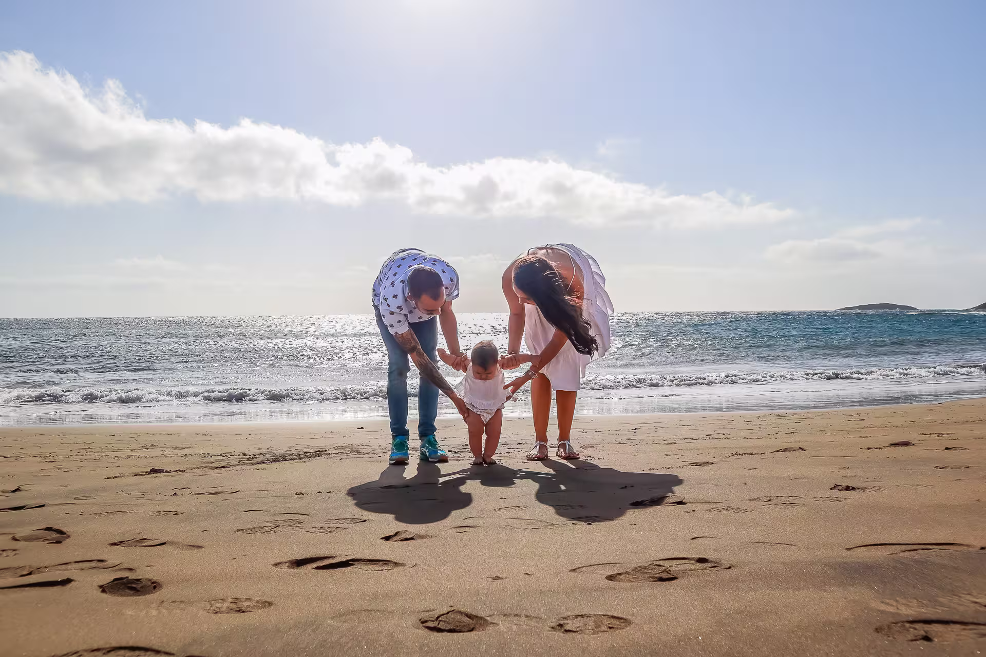 Eltern helfen ihrem Baby bei den ersten Schritten am weichen Sandstrand von Gran Canaria, mit dem Meer im Hintergrund. Einzigartiges Familien Fotoshooting Gran Canaria für Meilensteine
