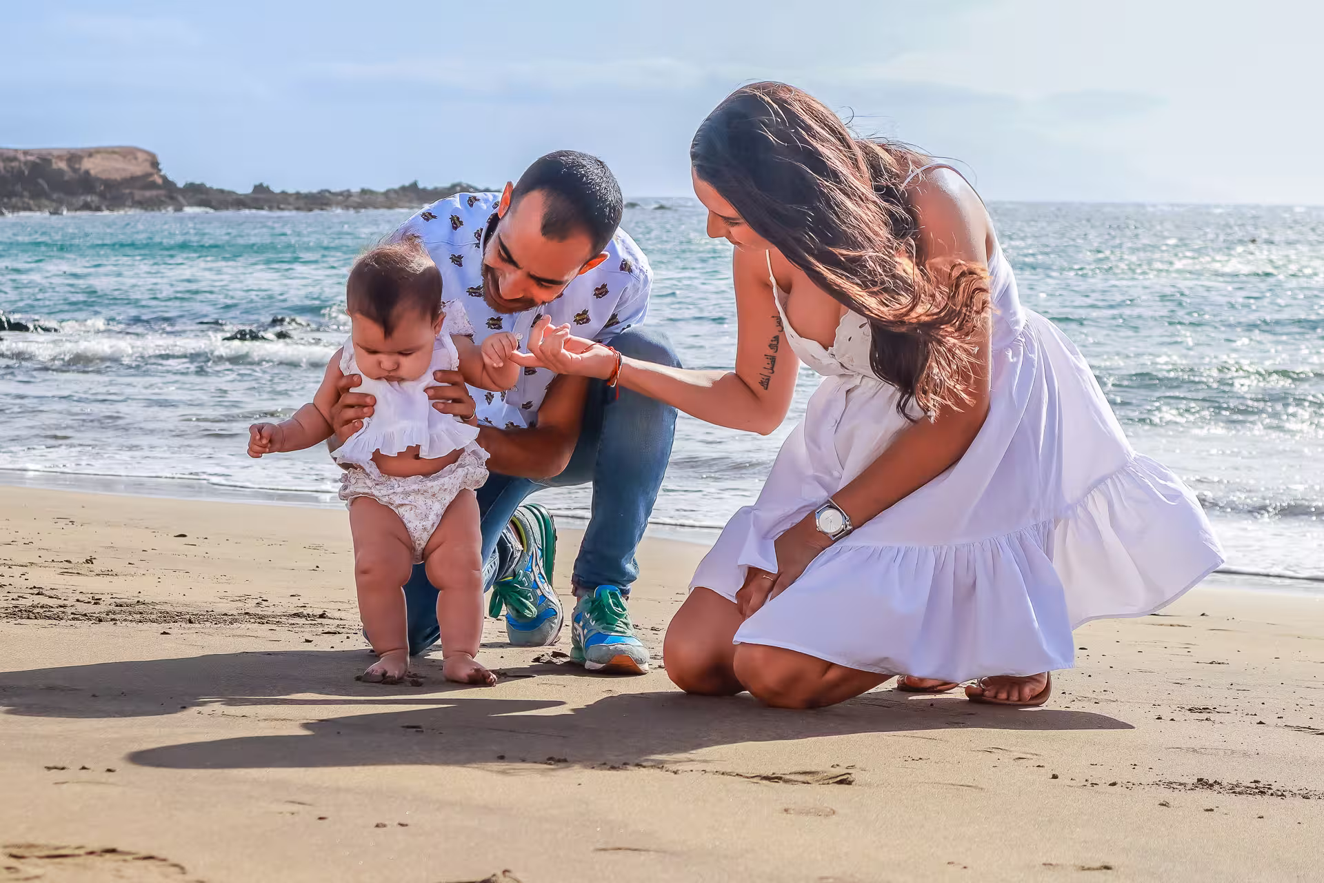 Eltern knien am Sandstrand von Gran Canaria und halten die Hände ihres Babys, das versucht, alleine zu stehen. Wertvolle Erinnerungen beim Familien Fotoshooting Gran Canaria.