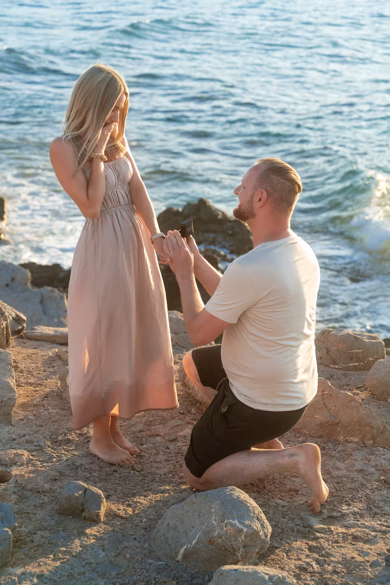 Emotionaler Heiratsantrag an einem felsigen Küstenabschnitt auf Mallorca. Der Mann kniet mit einem Ring vor seiner überraschten und gerührten Partnerin, mit dem Meer im Hintergrund