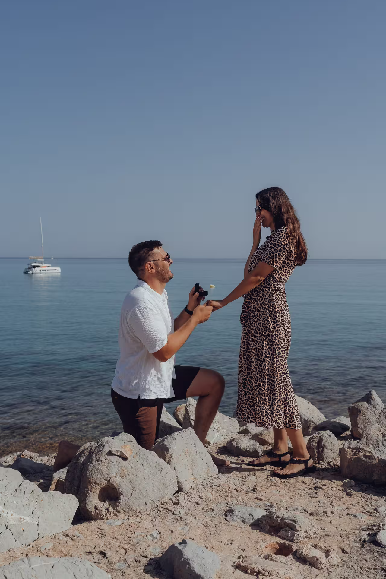 Heiratsantrag an einem sonnigen Tag an einer felsigen Bucht auf Mallorca. Der Mann kniet mit dem Ring vor seiner Partnerin, die erfreut die Hände vors Gesicht schlägt. Romantik am Mittelmeer