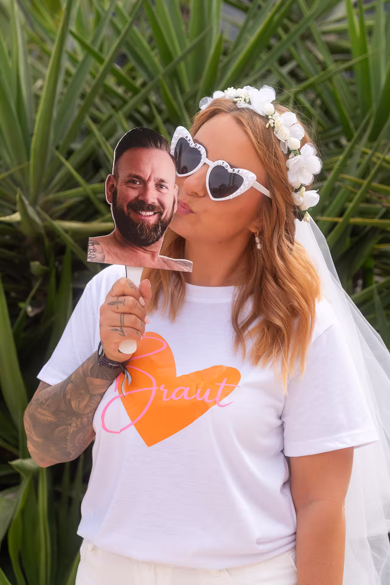 Fotoshooting JGA Mallorca: Die Braut gibt einem Foto des Bräutigams auf einem Stab einen Kuss, ein lustiges Accessoire für den Junggesellinnenabschied.