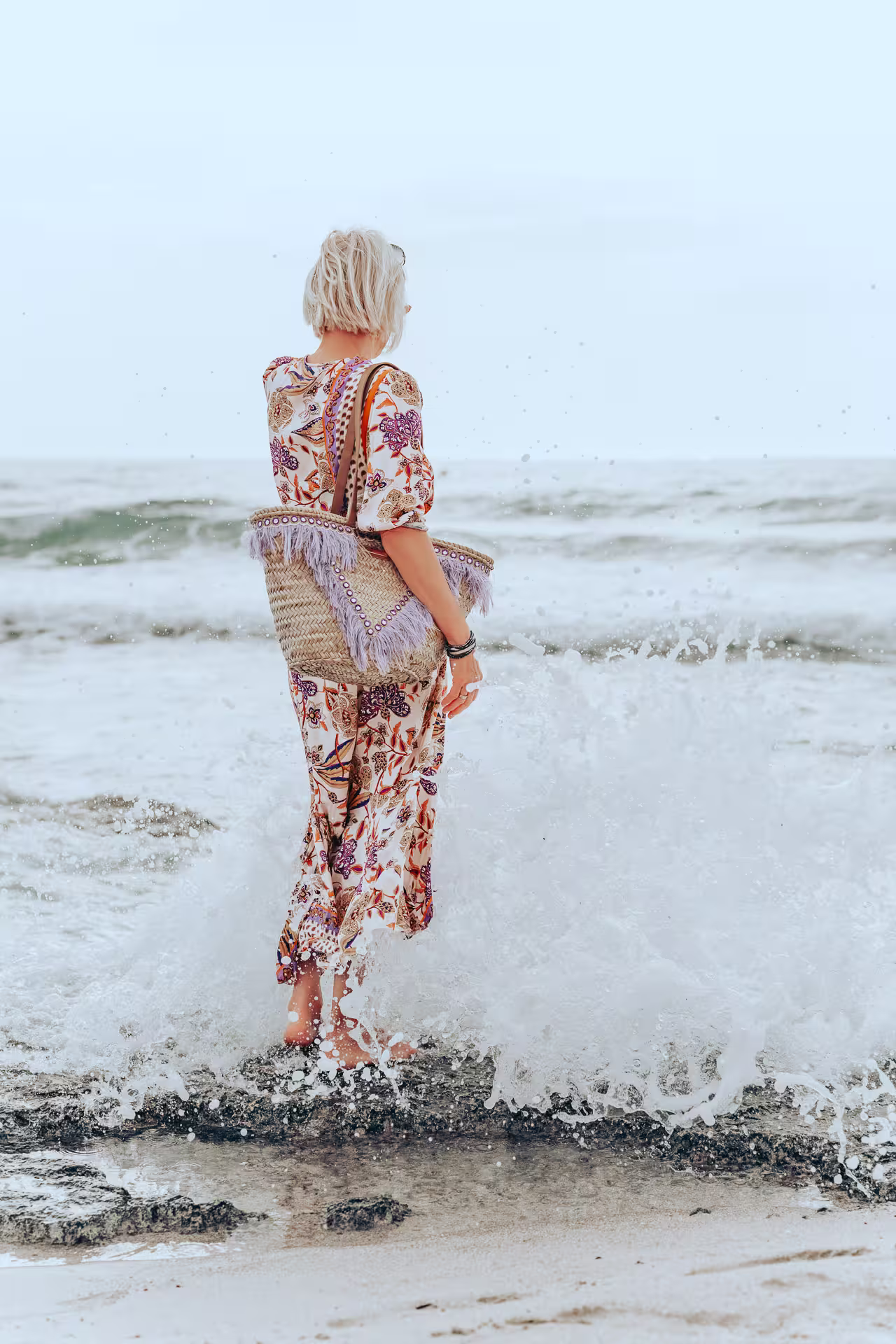 Elegante Frau in einem bunten Sommerkleid mit Korbtasche steht im flachen Meerwasser am Strand der Playa de Muro, während eine Welle um ihre Füße schlägt. Strandmode und Entspannung