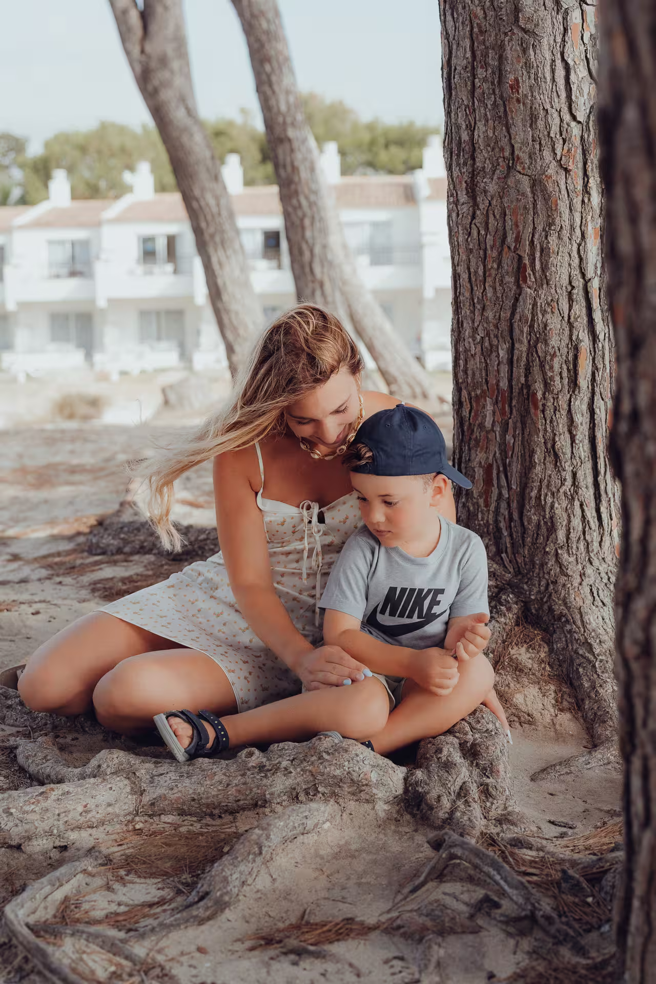 Junge Mutter mit wehendem Haar kniet neben ihrem kleinen Sohn im Schatten eines Baumes am Strand der Playa de Muro. Vertrauter Moment am Strand unter Pinien auf Mallorca