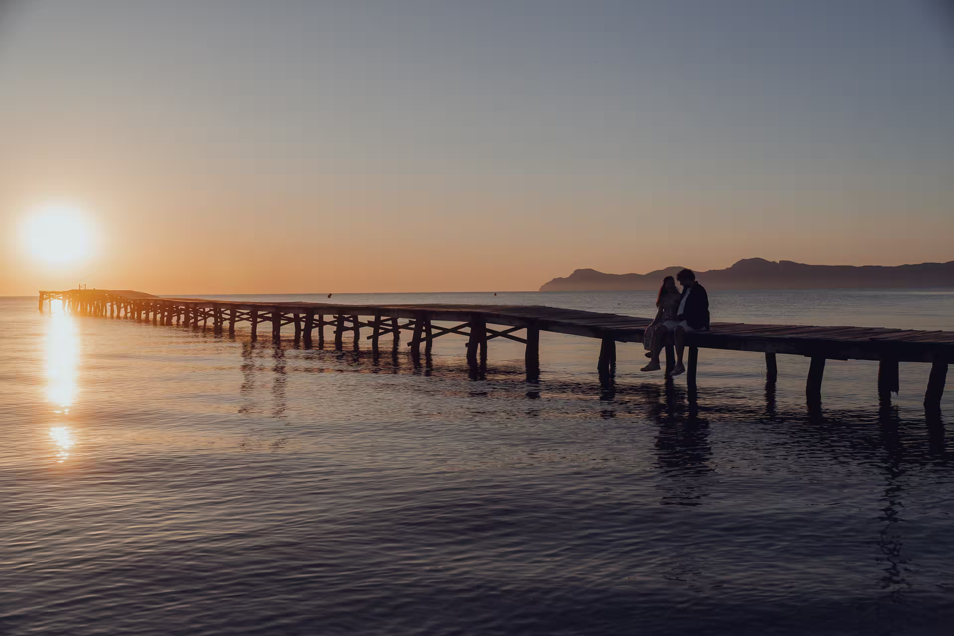Paar sitzt bei Sonnenaufgang auf einem langen Holzsteg, der ins Meer der Playa de Muro ragt, mit orangefarbenem Himmel und Bergen in der Ferne. Romantische Abendstimmung am Meer