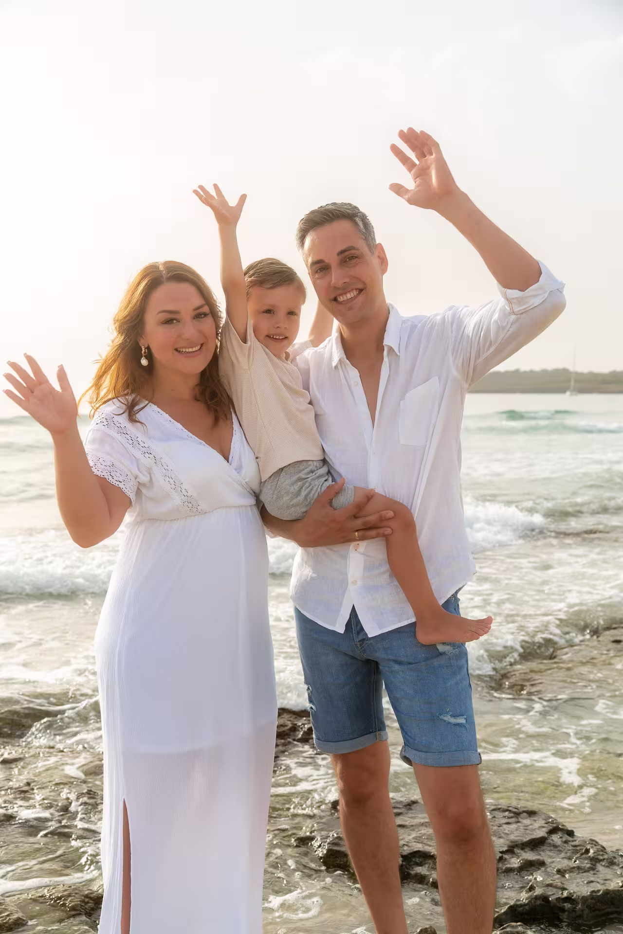 Fröhliche Familie, Vater hält den kleinen Sohn, winkt mit der schwangeren Mutter vom felsigen Ufer der Playa de Muro. Familienurlaub und Vorfreude auf das Baby am Meer.