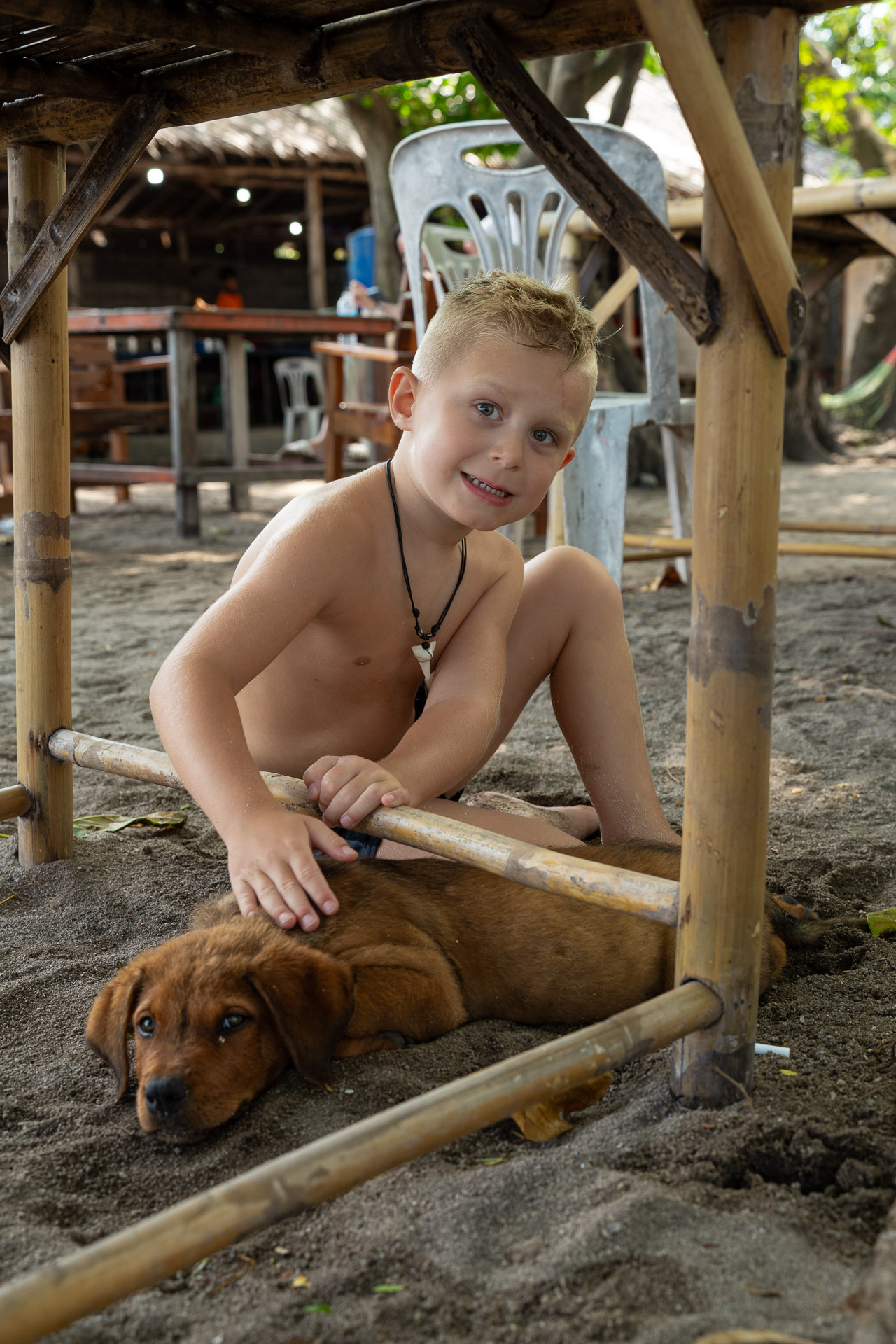 Kinderfoto in Thailand – Junge spielt im Sand und streichelt einen Hund, natürliche Urlaubsaufnahme vom Thailand Fotograf.