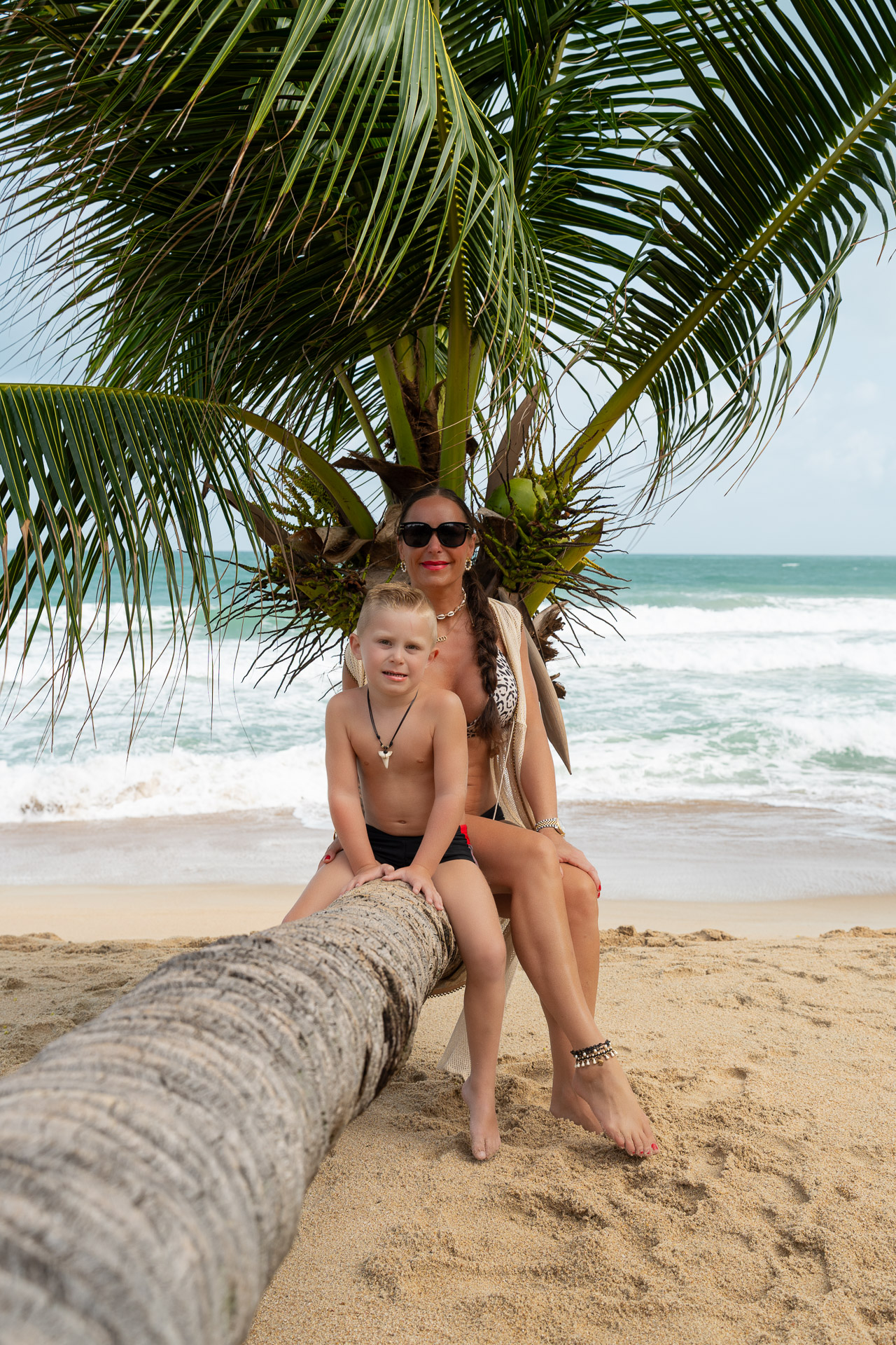 Familien Fotoshooting Thailand – Mutter mit Sohn auf umgestürzter Palme am Strand, natürliche Aufnahme mit Meerblick vom Thailand Fotograf.