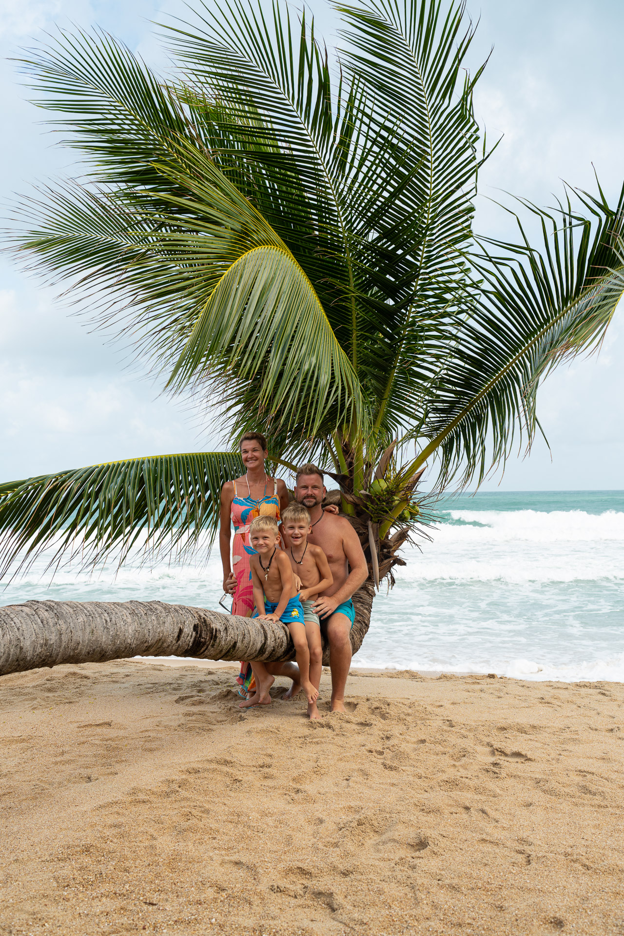 Familienfotoshooting in Thailand am Meer – Eltern und Kinder auf Palme am Strand, authentische Urlaubsaufnahme vom Thailand Fotograf.