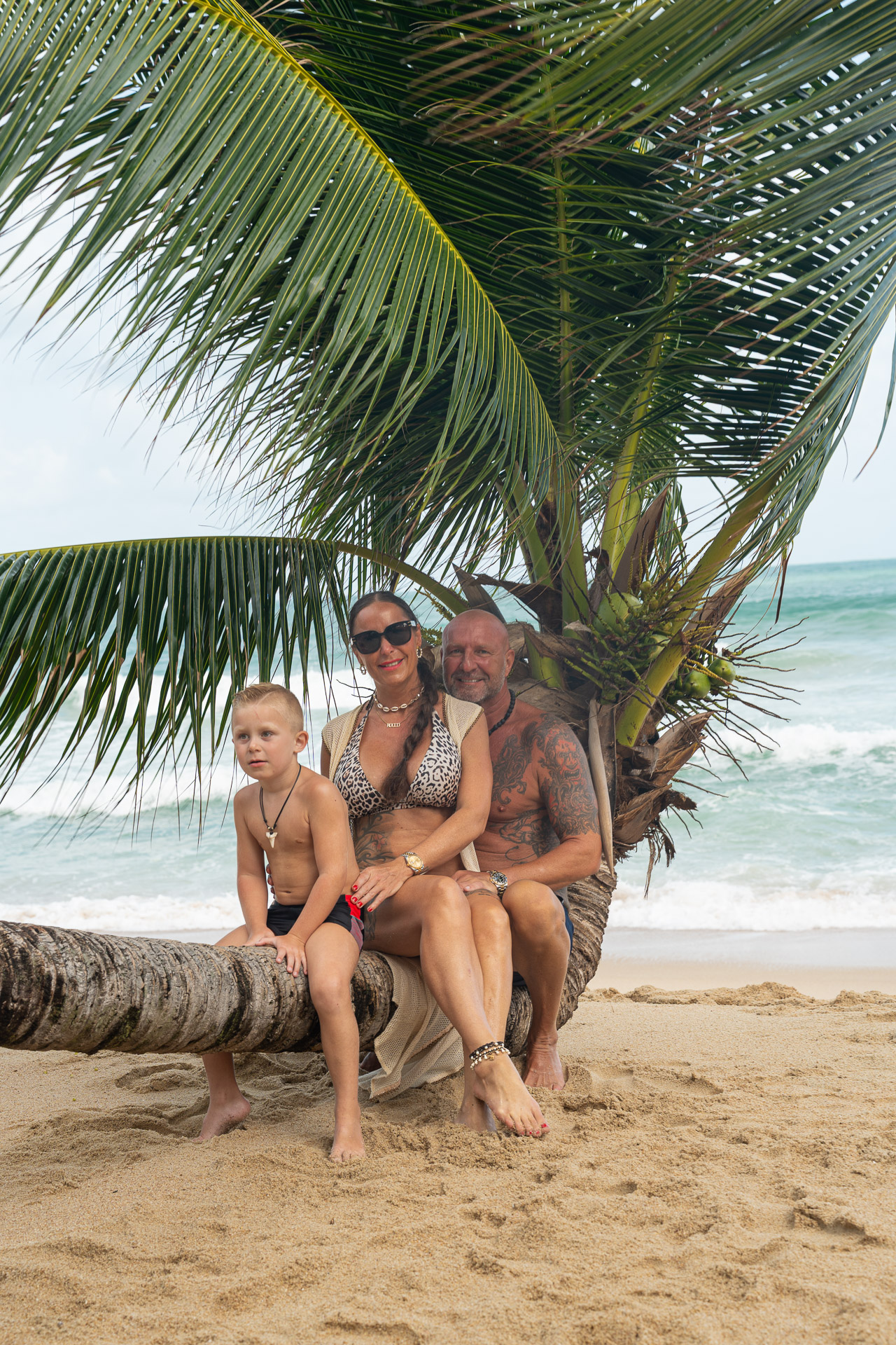 Familien Fotoshooting Thailand – Paar mit Sohn auf Palme am Strand, entspannte Urlaubsbilder unter Palmen, festgehalten vom Thailand Fotograf.