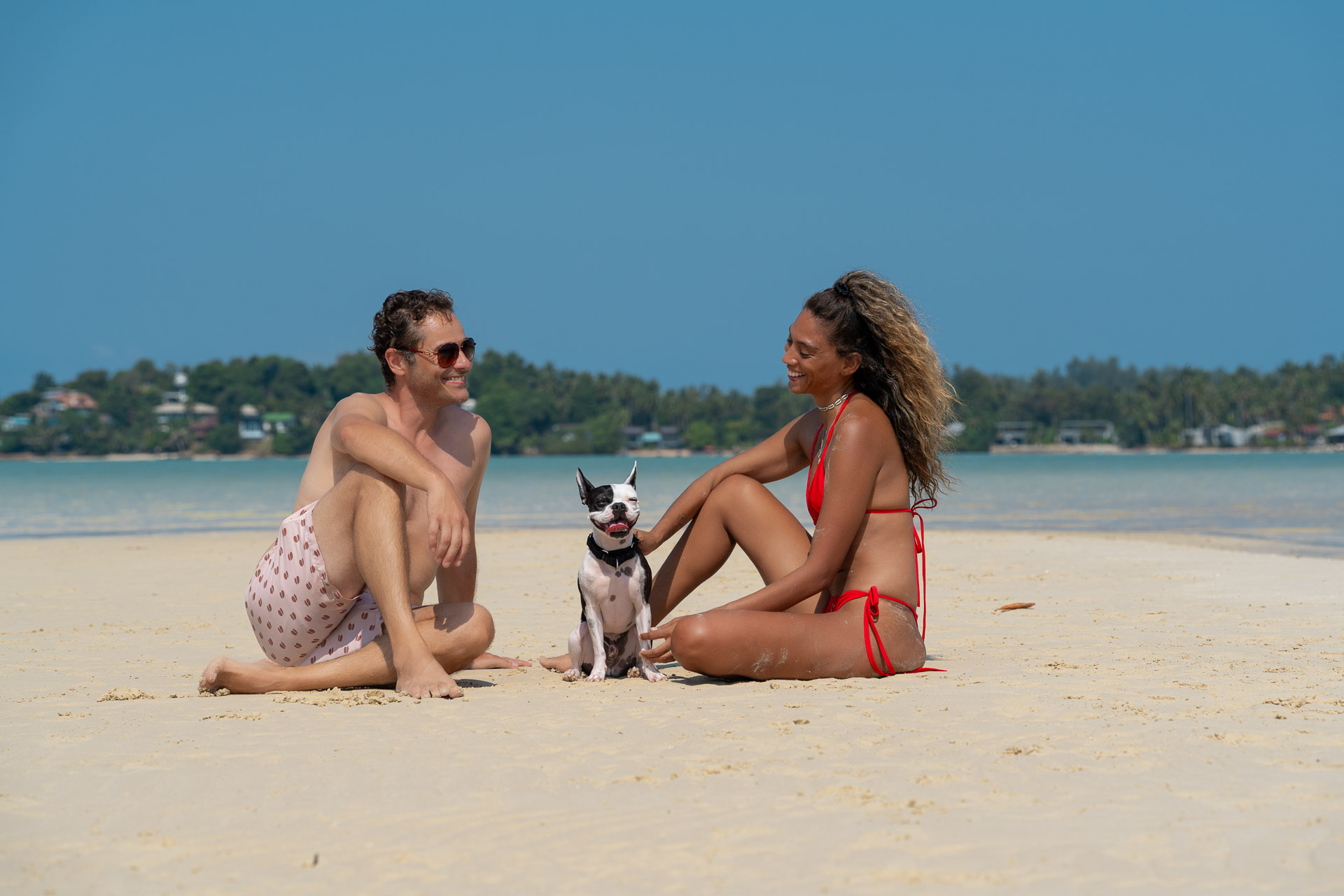 Paar Fotoshooting in Thailand am Strand – Mann und Frau sitzen entspannt im Sand mit Hund, natürliche Momentaufnahme vom Thailand Fotograf.